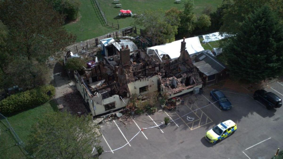 An aerial shot of the pub shows the building has been gutted. Only the foundations and part of the facade remain. A police car is parked in the car park as well as another two cars. To one side is a white marquee.