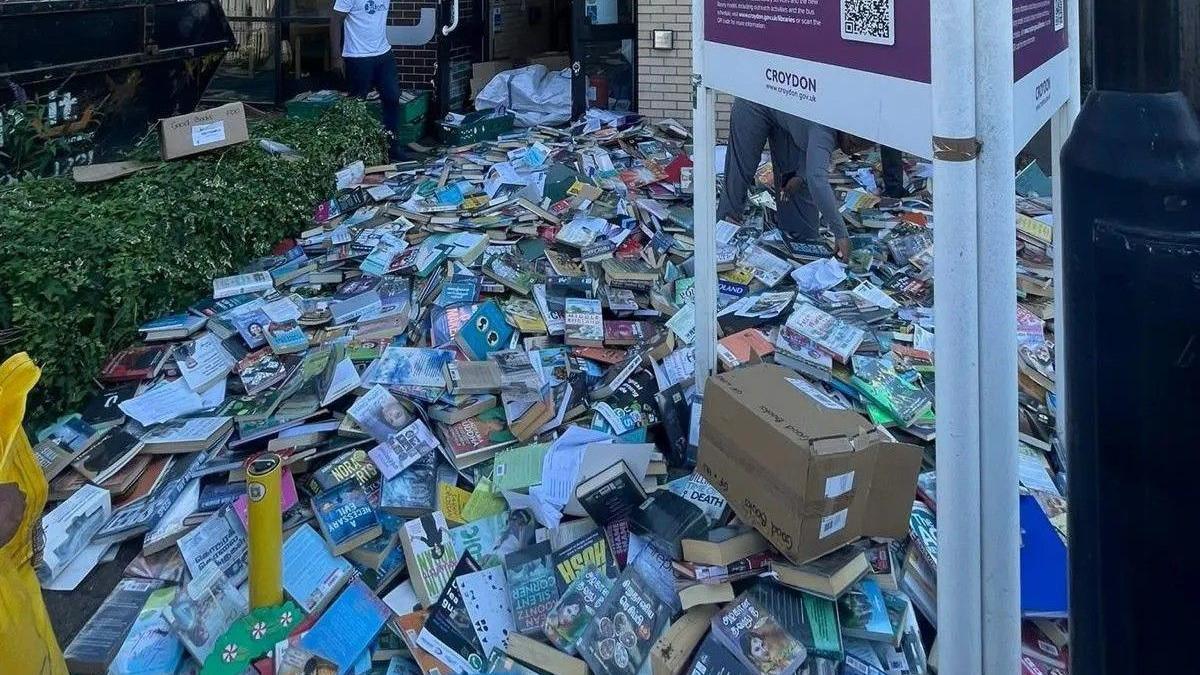 Piles of books cover the pavement outside Broad Green Library in Croydon, many in good condition and some in open boxes. Two people are visible among the stacks, with a black skip to the left and a library sign in the foreground.