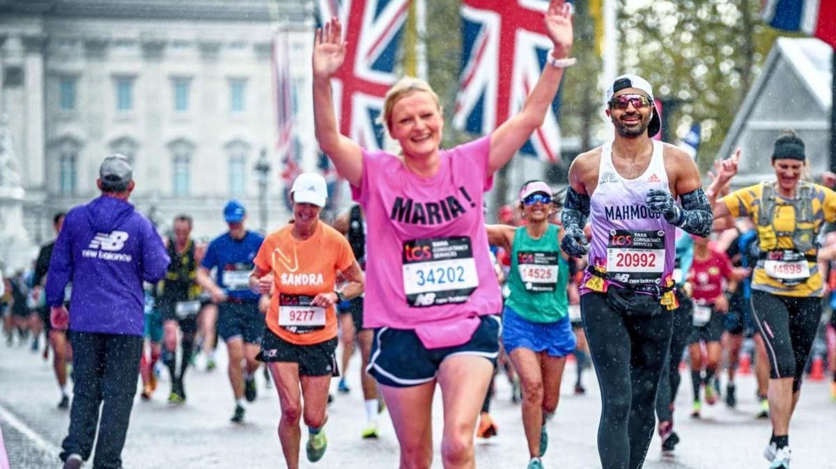 Maria Mulgrew running a competitive long-distance race.  She is wearing a bright pink T-shirt decorated with her name "Maria!" in black letters, and black running shorts with white seams.   She is smiling and has both arms raised above her head. 