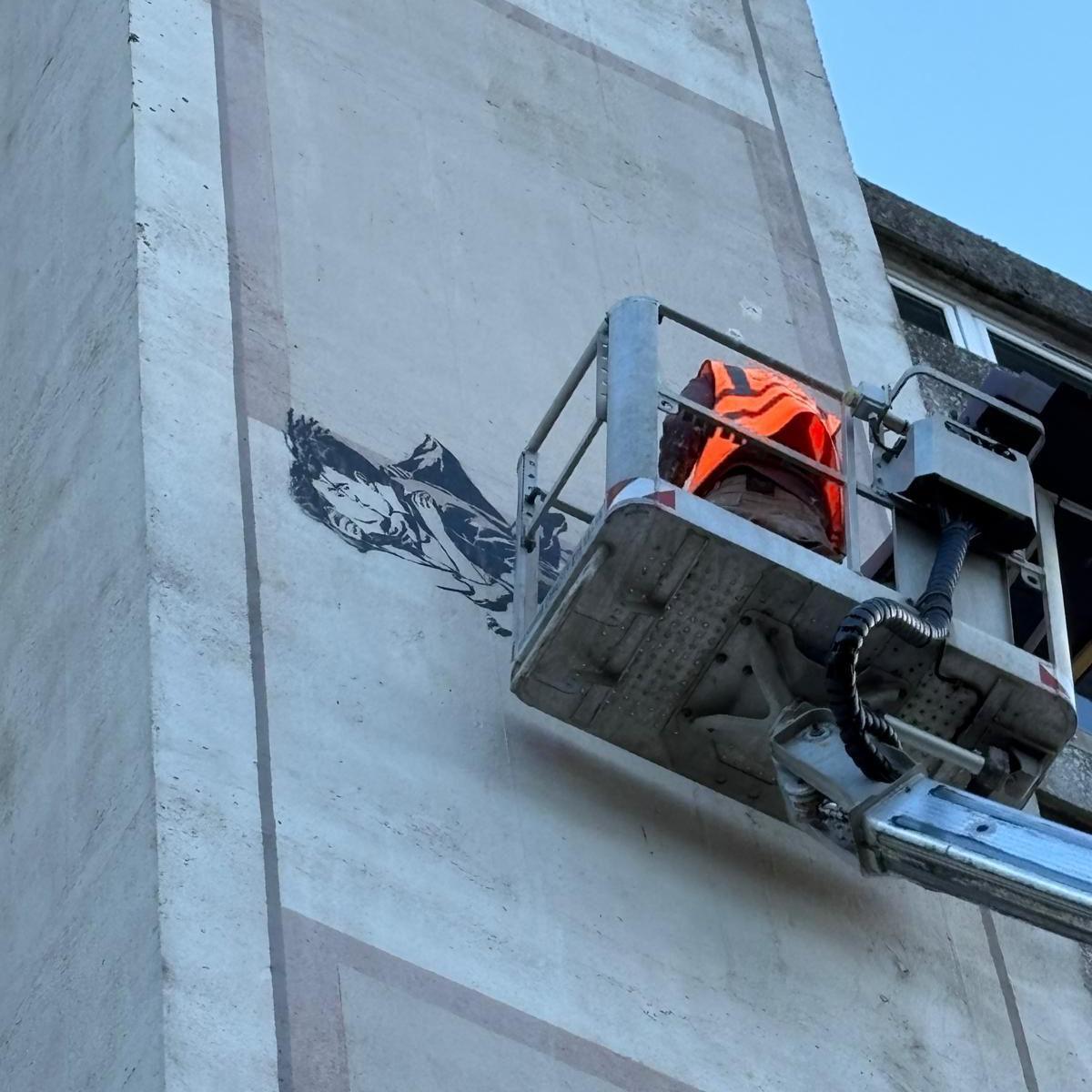 A man in an orange hi-viz jacket on top of a cherry picker platform painting a mural on the side of the building.