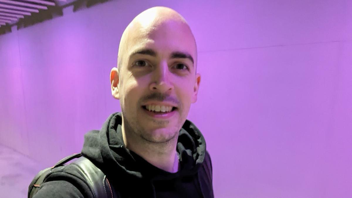 Dean Stokes - a man with no hair smiles into the camera. He has a black hoodie on and has a black bag over his shoulder.