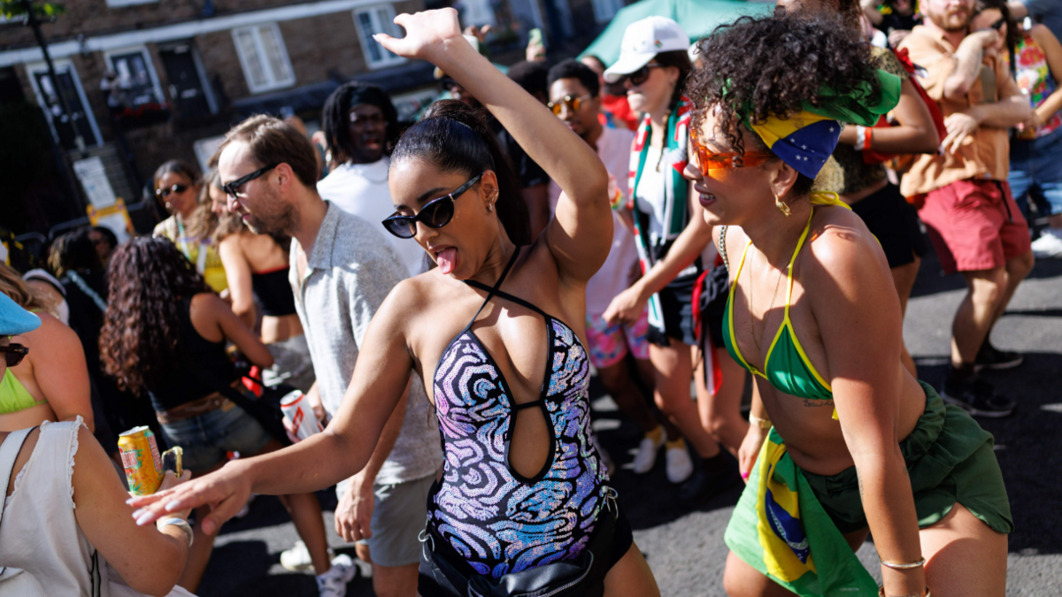 Two women dance in the street surrounded by other people in the crowd. One wears a sequin leotard and another wears a green and yellow bikini top and green shorts.