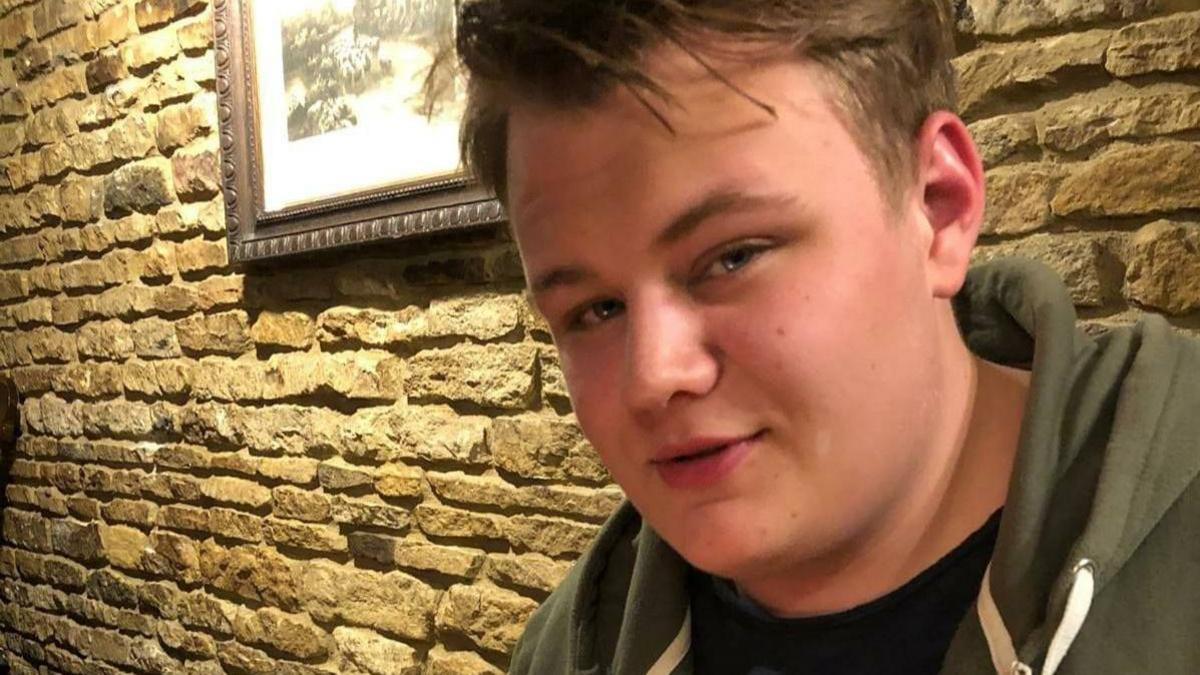 Harry Dunn had fair hair and was wearing a khaki green hoodie and black T-shirt. Behind him is a stone wall with a framed picture.