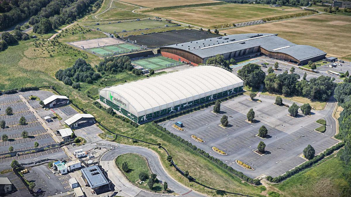 An aerial view of a large building with a grey roof, surrounded by car parks, sports pitches, and fields. In the centre of the shot is a CGI, large, white hall with a PadelStars sign on the side.
