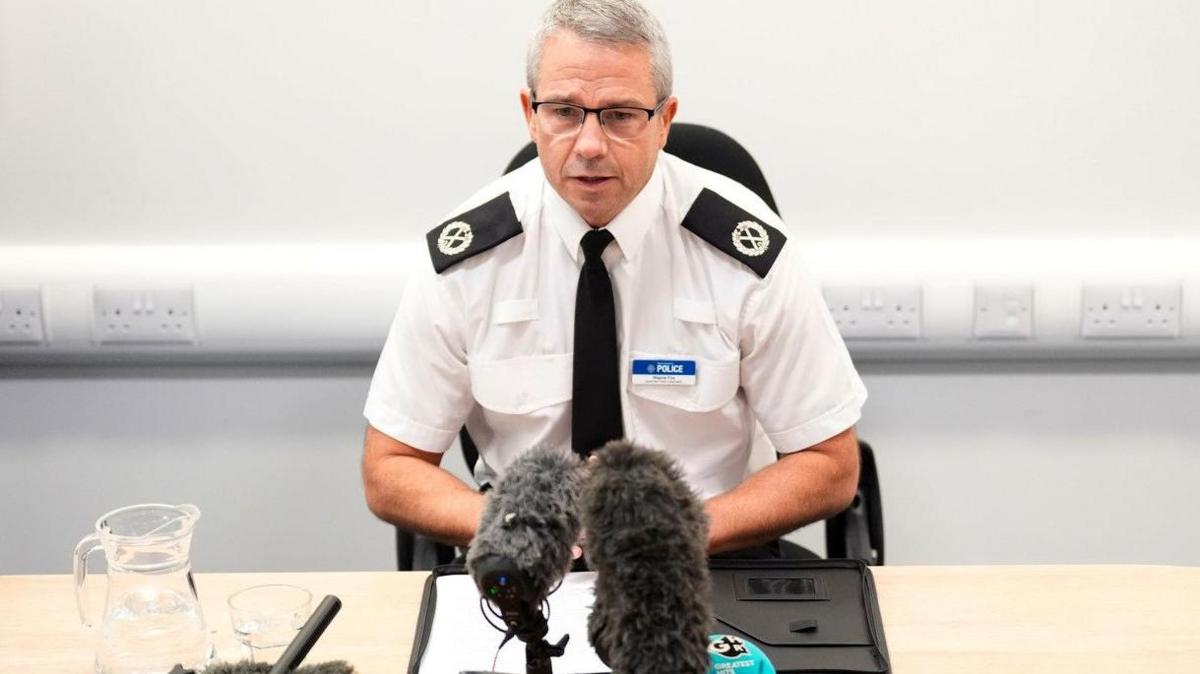 Picture of a man wearing a police uniform sat behind a desk with a microphone in front of him