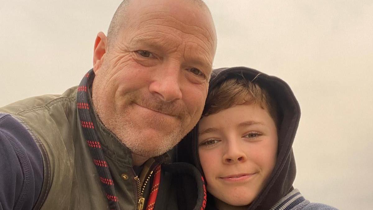 A selfie of a bald ban in a green and blue jacket next to a boy wearing a black hoodie. Both are looking into the camera and smiling