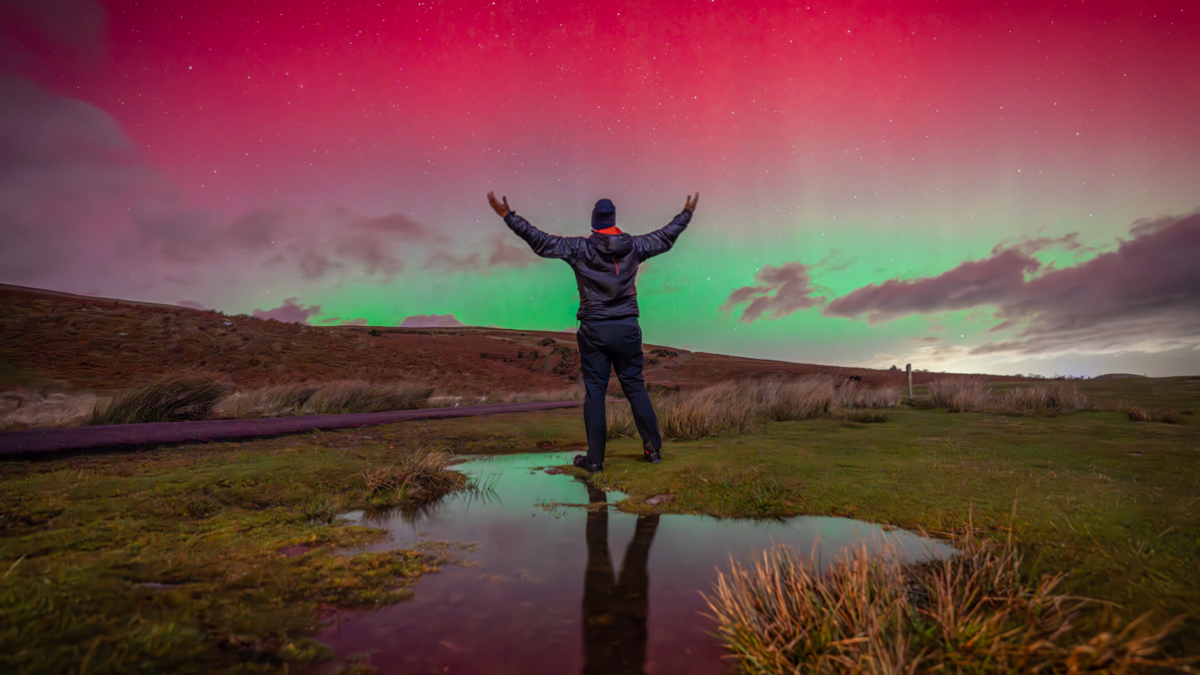 A person wearing a navy coat and woollen hat stands with their arms wide - looking at the pink and green Northern Lights in the sky. The glow reflects in a puddle on the ground.