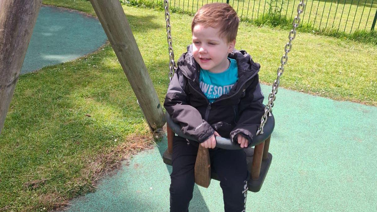 A young boy with short brown hair, a light blue shirt, a black coat and dark trousers, sits in a swing at a playground.