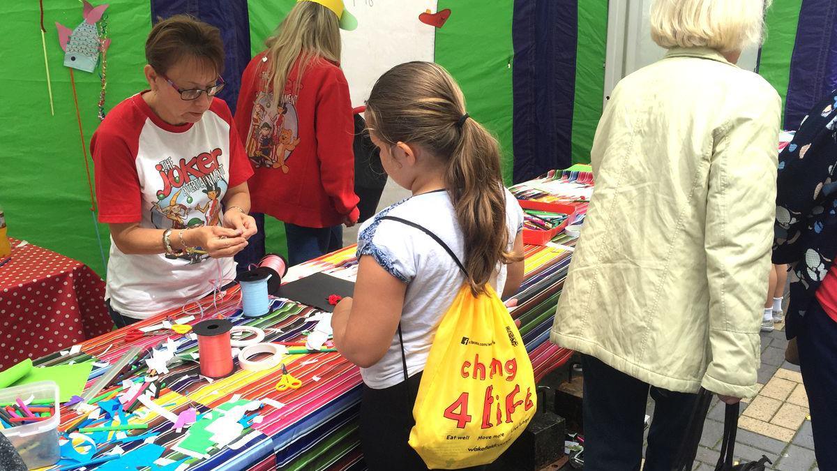 A child wearing a white t-shirt and a yellow 'change 4 life' drawstring bag standing in front of a craft materials stall with a colourful tablecloth. A woman with a 'the joker' white and red t-shit is stood behind the stall.