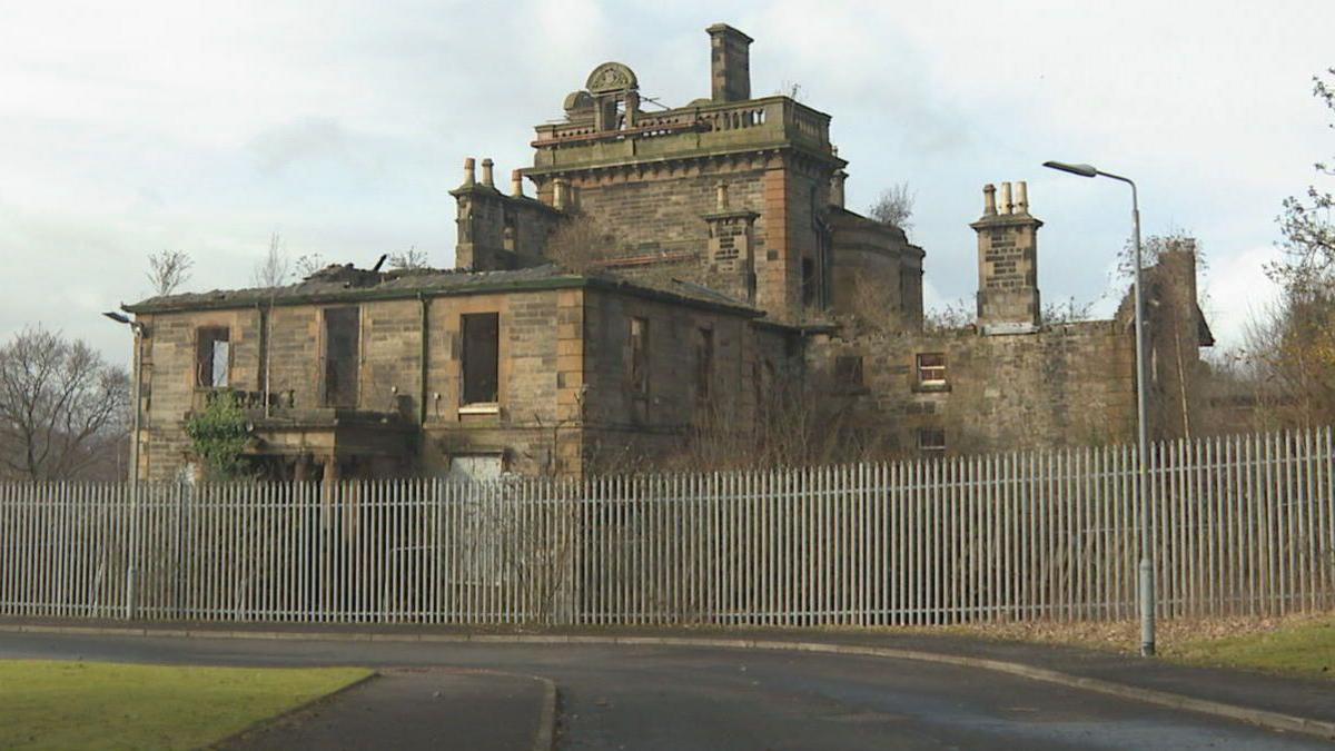 A dilapidated school building behind a metal fence - parts of the roof is missing.