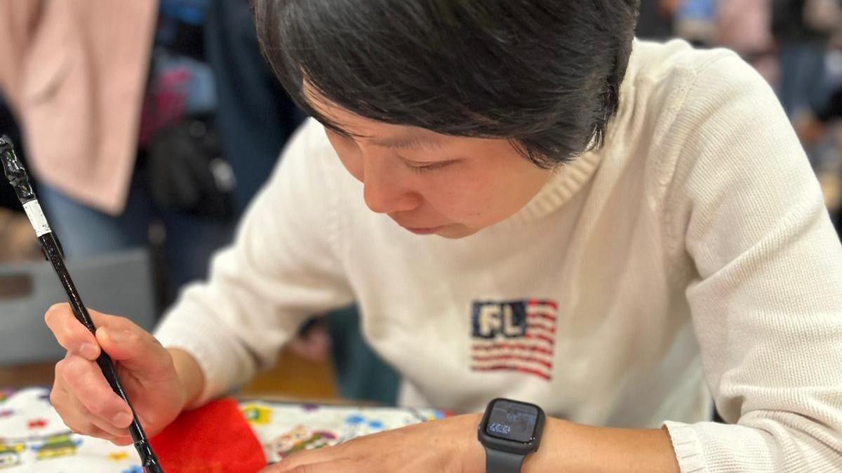 A woman in a cream sweatshirt with a red white and blue flag. She is bent over a table and holds a black paintbrush.  She has black short hair swept across her face.