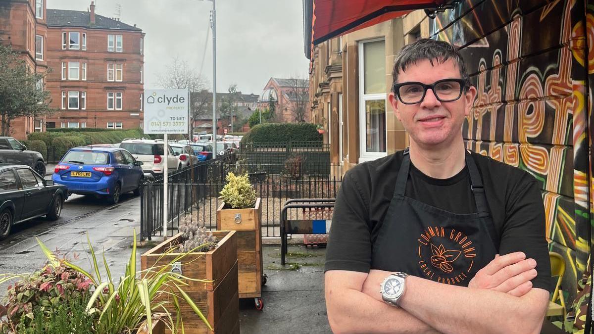 A man with dark glasses stands outside a coffee shop smiling.