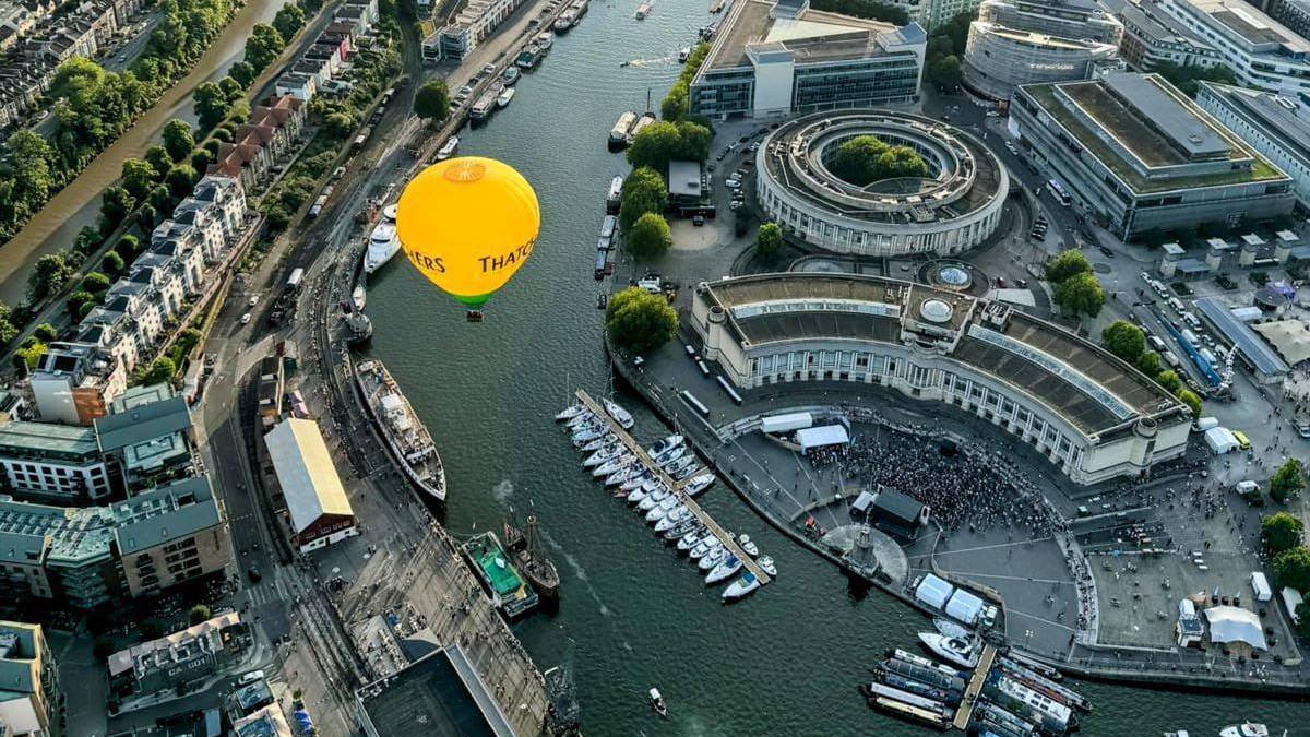 An aerial view looking down towards Bristol Harbour and Lloyds Amphitheatre. There are lots of boats moored in the harbour and curved buildings beside the river. A yellow Thatcher's hot air balloon is floating above the water.  