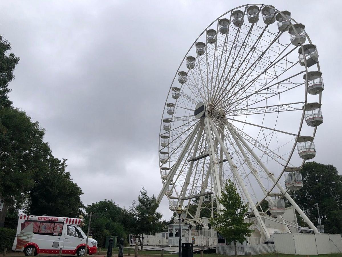  A big ferris wheel, white, with several carriages hung around the wheel while a red and white coloured ice cream van stands in front.