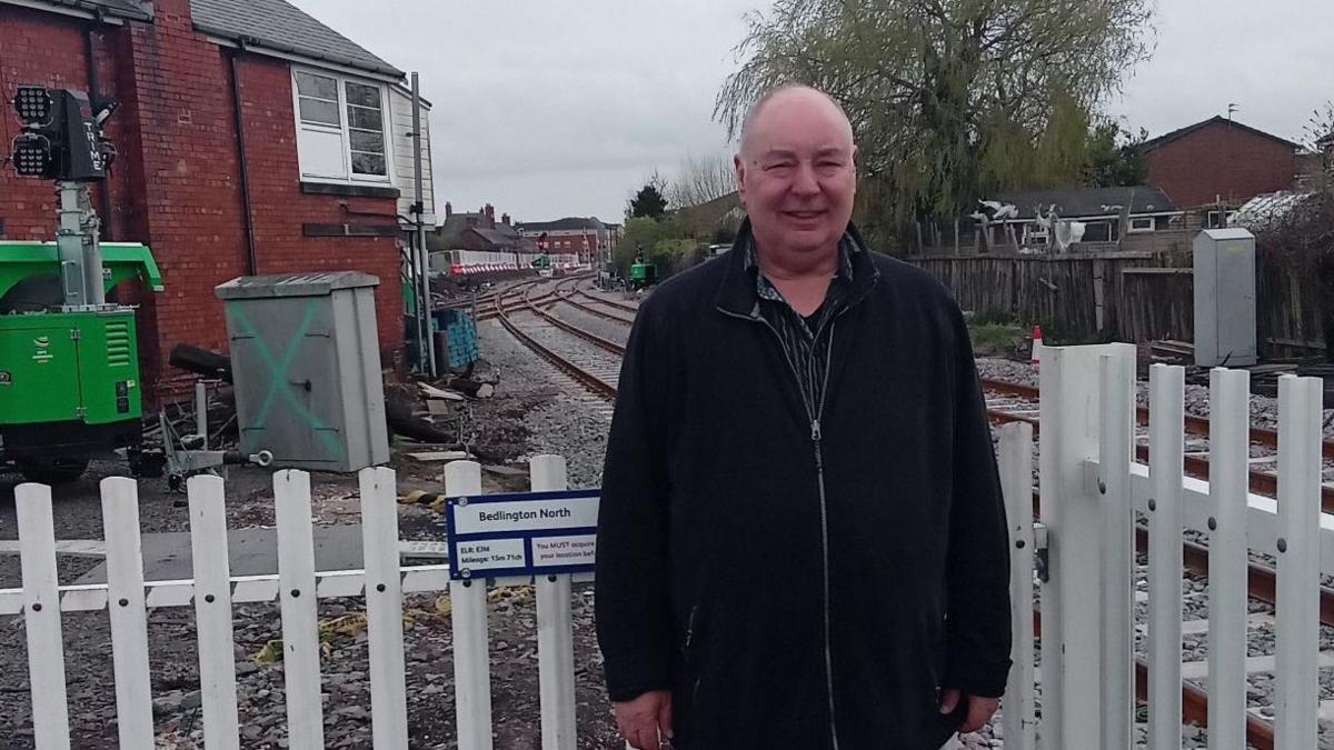 Dennis Fancett is standing in front of a barrier at Bedlington Station on the Northumberland Line. You can see a barrier behind him