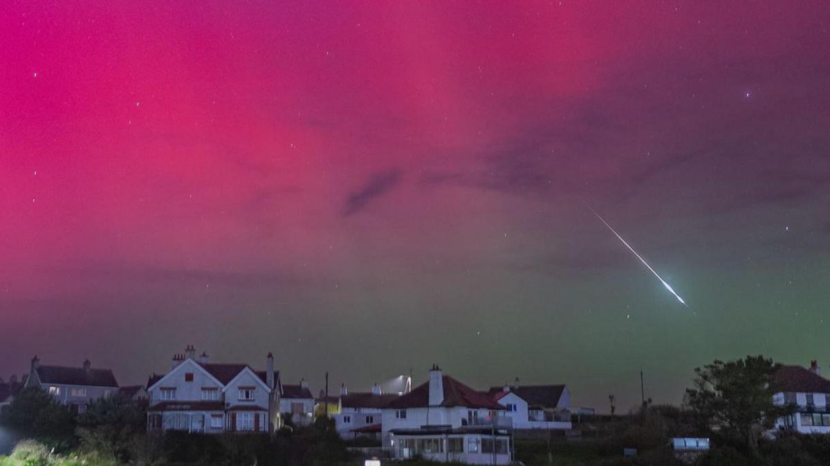 A meteor in the purple/pink sky. There are white houses with red tile roofs in the foreground of the picture.