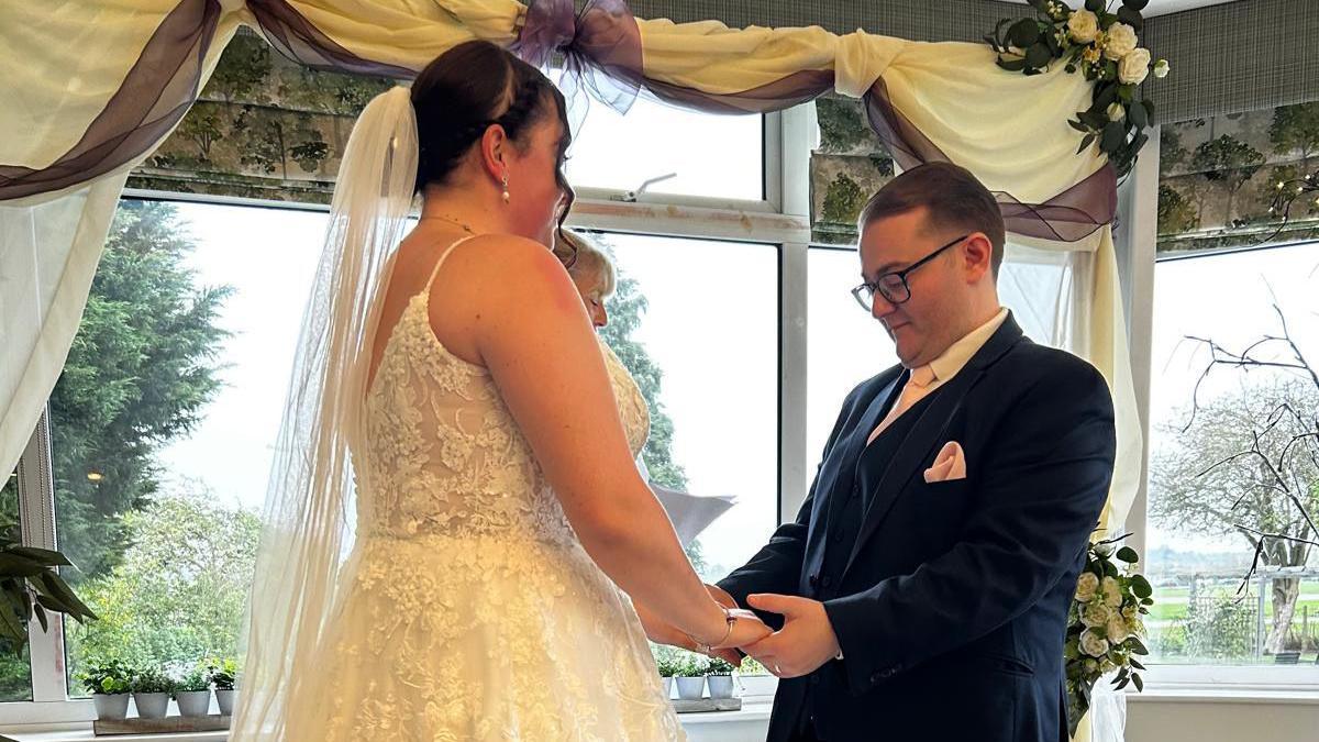 Lauren and Jack are pictured during their marriage blessing ceremony. They are facing each other and holding hands. Lauren is in her white wedding dress with a long veil placed behind her head. Jack is wearing a navy suit with a white shirt. He has short dark hair and is wearing black glasses.