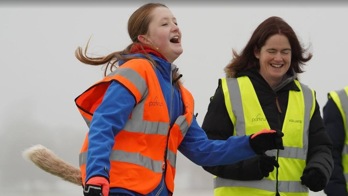 Josie in a role as a "tail" walker for York parkrun. She has long red hair tied back in a ponytail and  wears a bright orange reflective vest with the parkrun logo and a fake fox tail. 