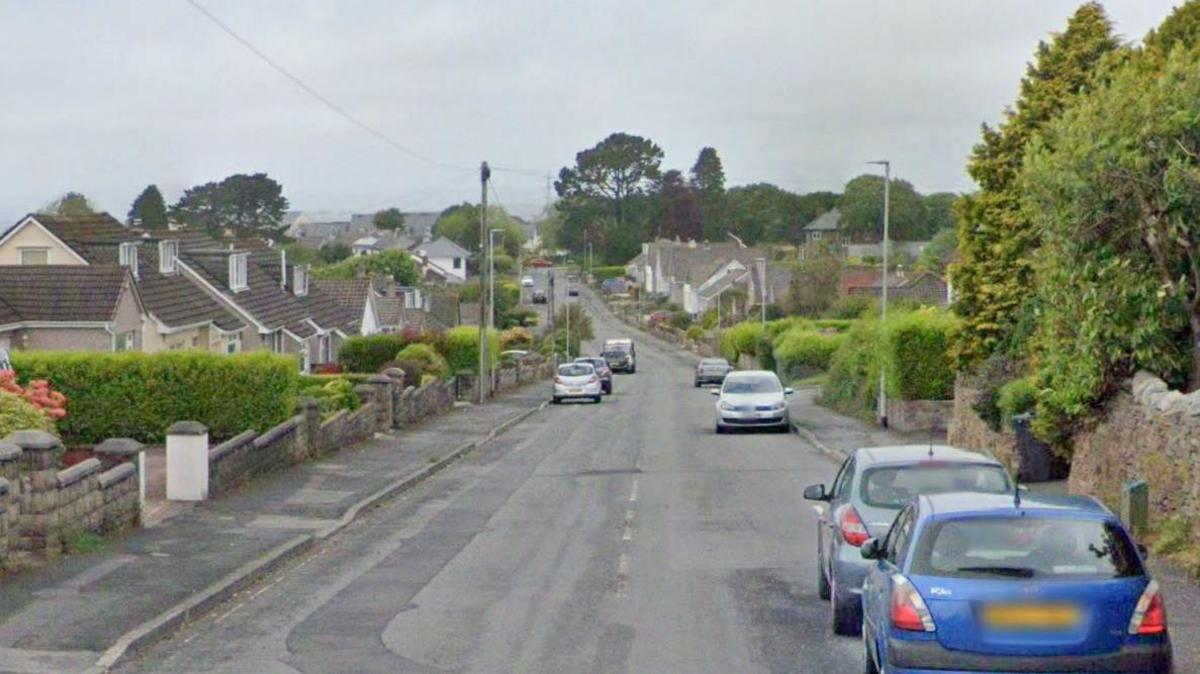 A view down the lane. It is a residential street with cars parked on either side of the roads.