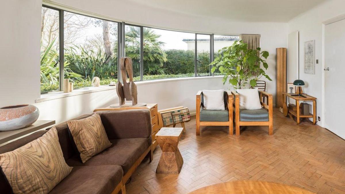 A large curved window dominates the white-pained interior, which has a herringbone wooden floor and a wooden-framed sofa, chairs and tables.