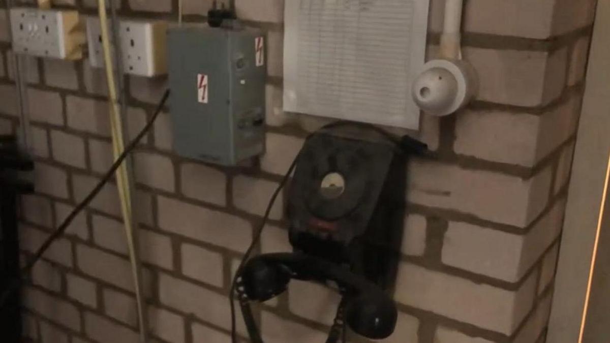 Black rotary-dial phone with receiver hung below it. There is a white checklist above it. There is also a metal electrical circuit box alongside and power sockets. They are all mounted on a brick wall.