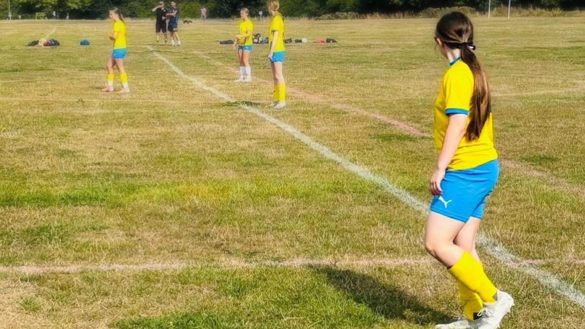 Several young girls in yellow football tops and blue shorts on a grassy football pitch.
