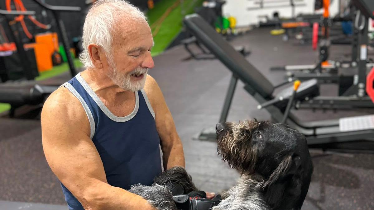 Mick Wadge - a white-haired man in a vest top, surrounded by gym equipment - smiles at a black and grey dog standing on its hind legs