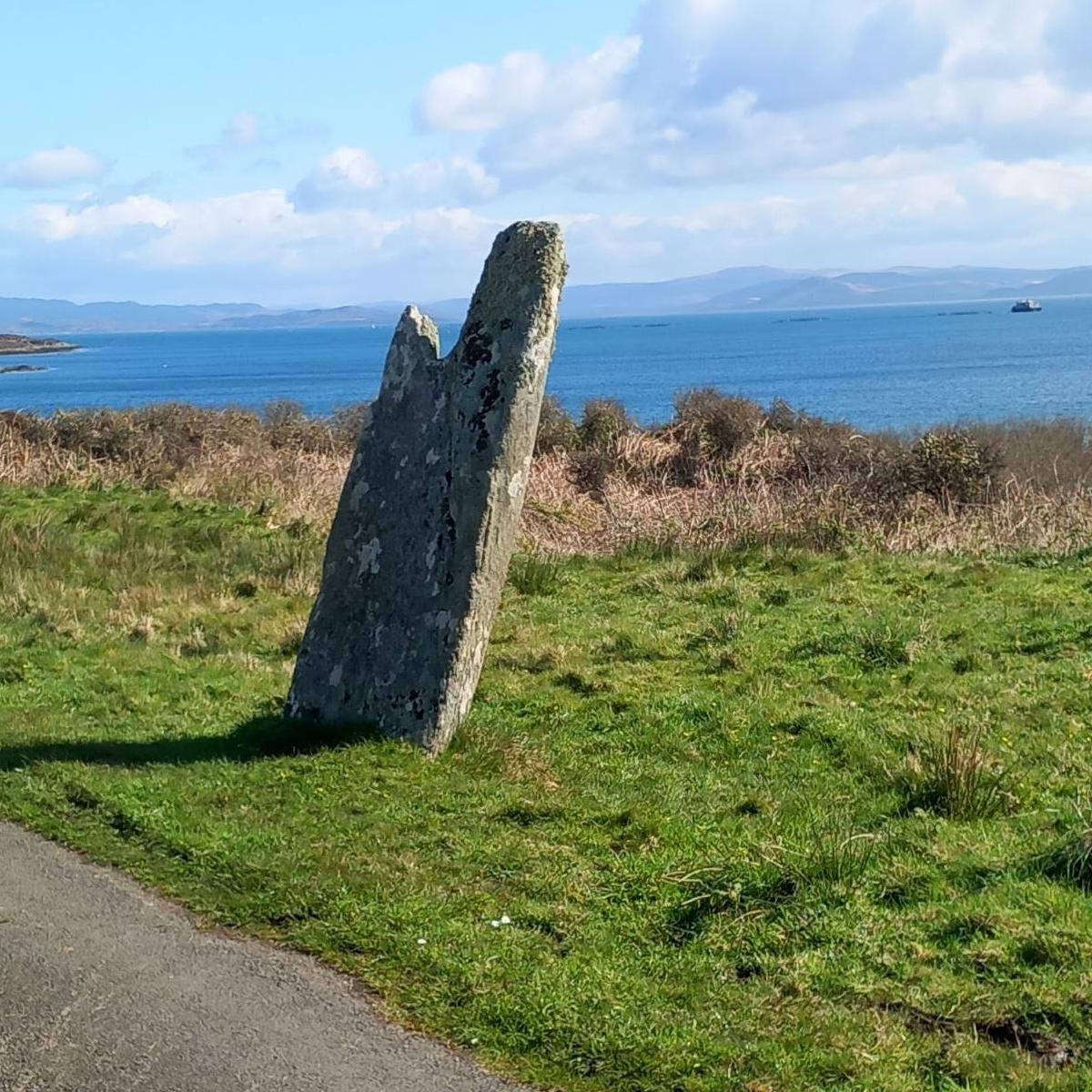 A standing stone, leaning to one side, near the sea on the island of Gigha. There is a boat in the distance.