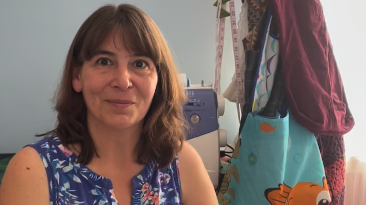 Sharon Goold smiles at the camera - she wears a blue floral top and has a sewing machine behind her. Hanging to the right are some colourful fabric tote bags