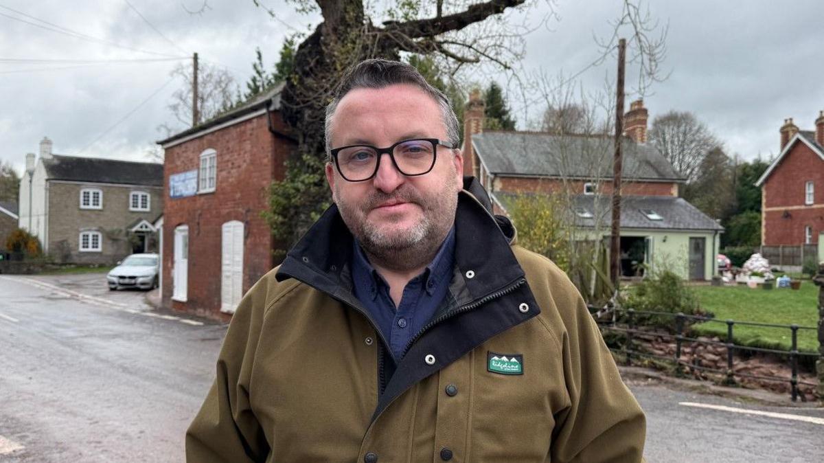 A man wearing glasses in a brown coat and blue shirt. Homes and a road are behind him.