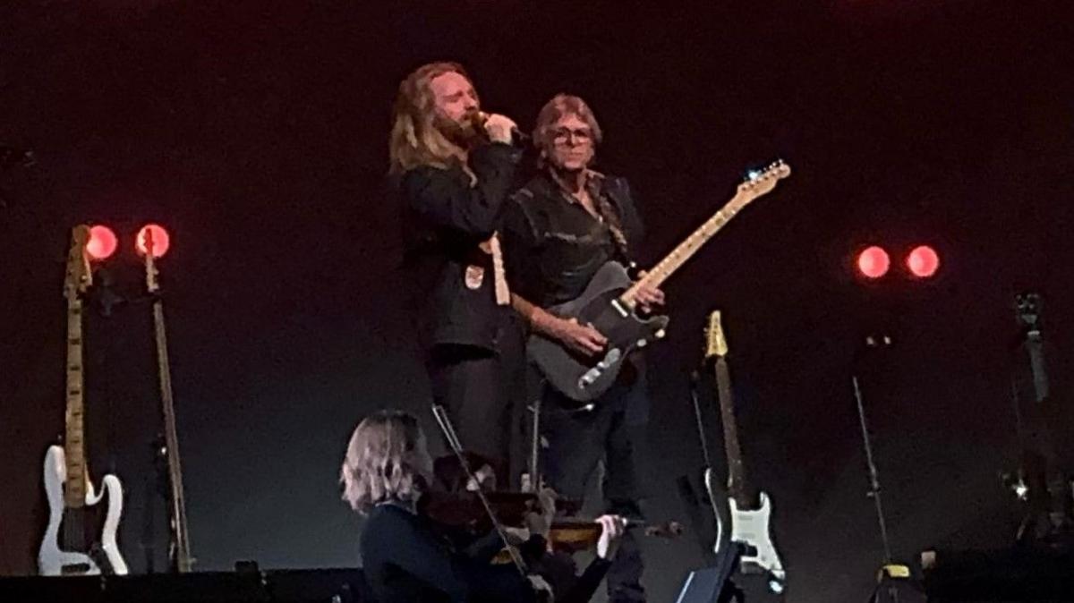 Kelvin Pratt stands on stage, dressed in black, playing a black guitar. Next to him is Sam Ryder, also wearing black, with long hair. He is holding a microphone to his mouth. Several guitars can be seen on stands nearby.