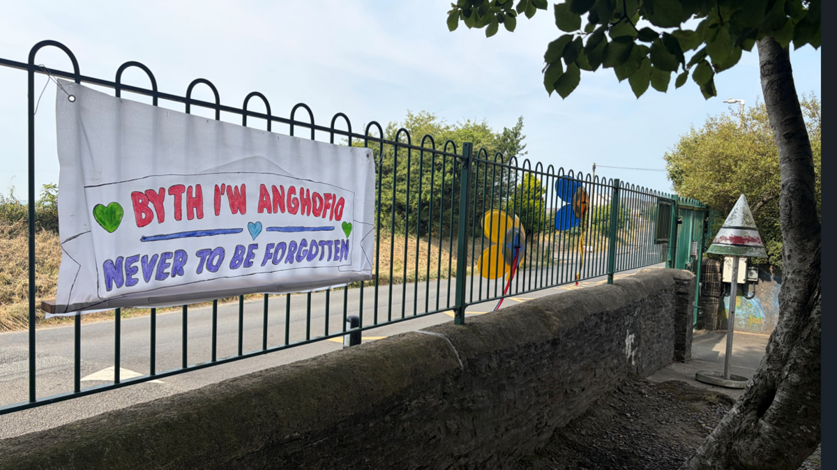 Ffens ysgol gyda baner mawr arno'n dweud 'Byth i'w anghofio - Never to be forgotten'