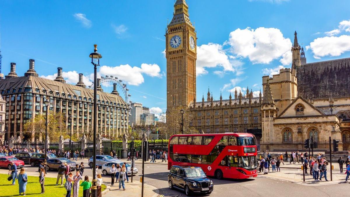 A view of the UK Parliament with the Elizabeth Tower in the background, a red double-decker bus, and several people walking near the grass area.