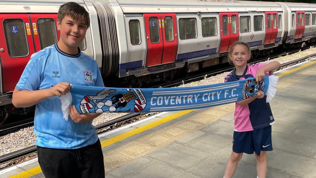Dexter with his sister Sophia at Coventry City's play off final at Wembley in London. They are a tube station, both wearing Coventry City shirts. They are holding either side of a Coventry City scarf. There is an iconic London Underground train behind them.