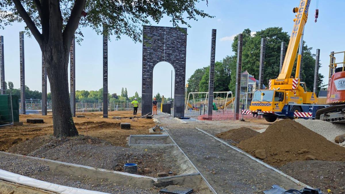 A large number of brick and slate looking heritage poles, on a construction site. There are paths, being built, with grey slate, large mechanical machinery to the right, and a large tree to the left. Workers are on the site.