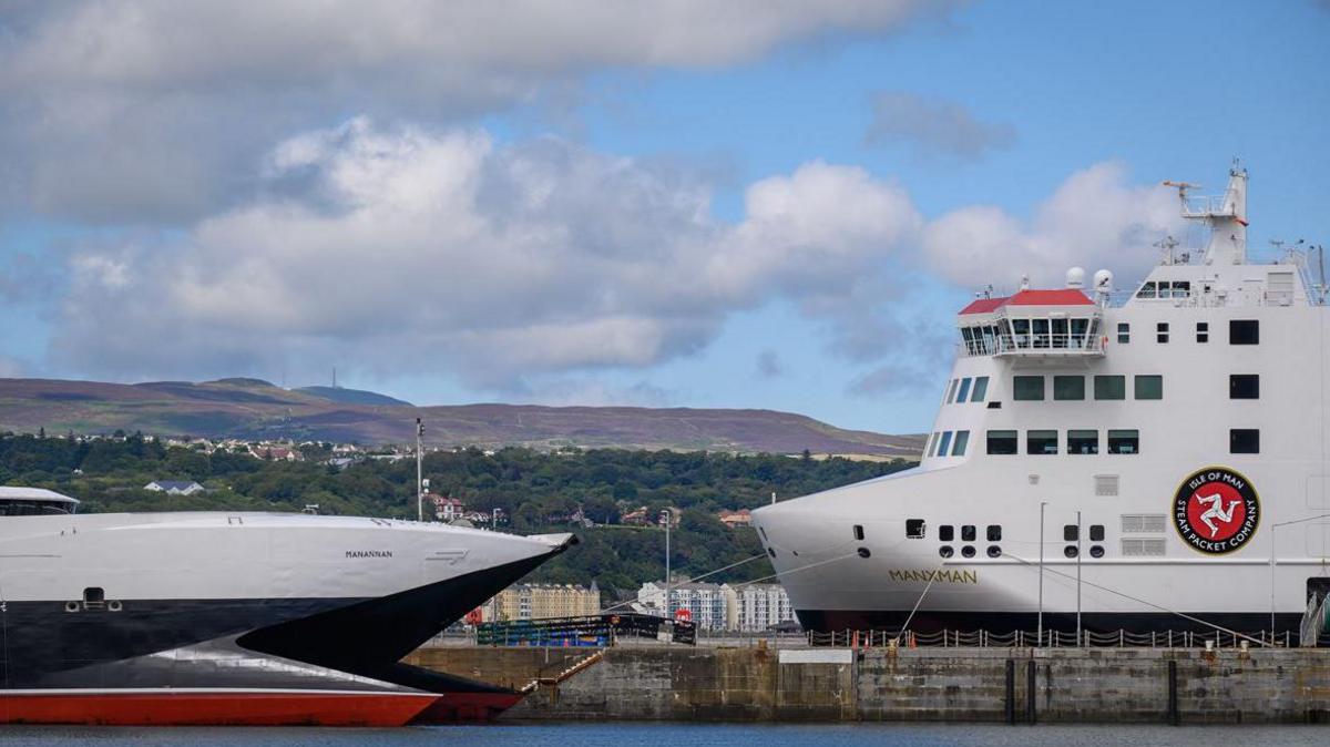 The Manxman ferry docked in Douglas Harbour next the Manannan. The ferries are white, black and red. There are green hills in the background and a blue sky with white clouds.