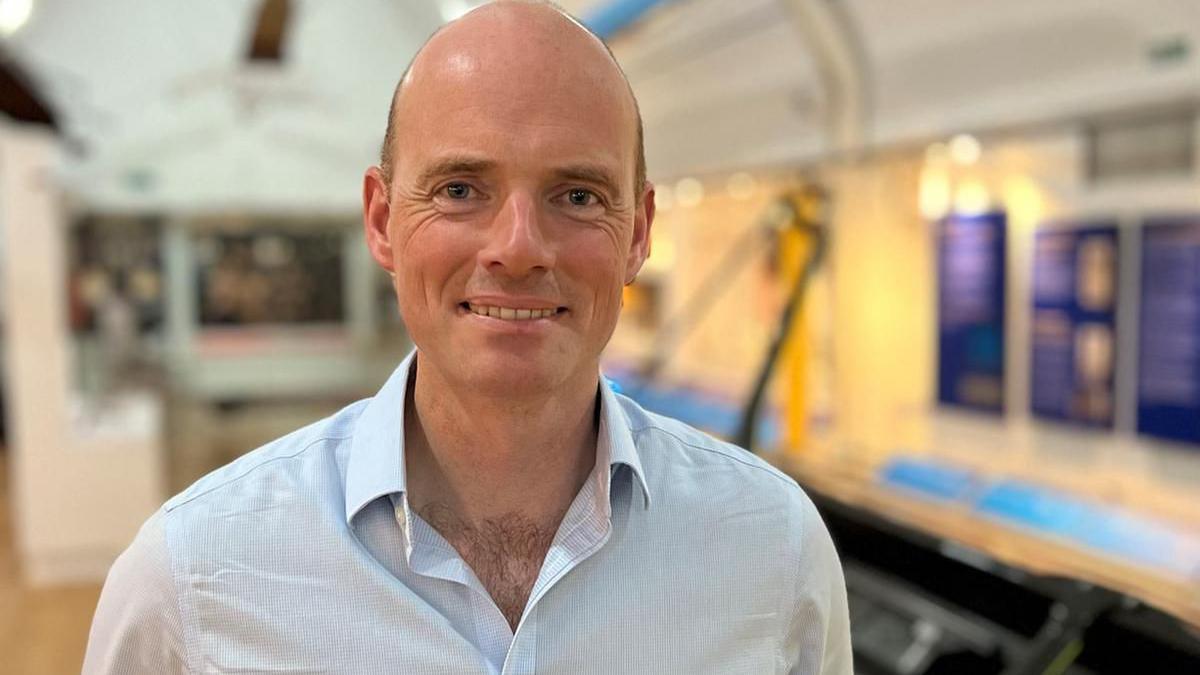 A bald man wearing a white shirt, smiling and looking at the camera. Museum exhibits can be seen in soft focus behind him.