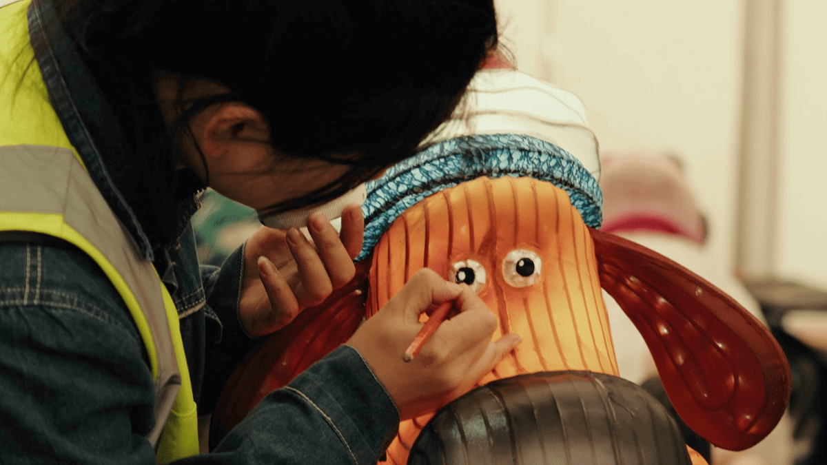 A sculpture of Gromit with a woolly hat, from the TV series of Wallace and Gromit. A woman in a high-vis jacket is using a small pencil to add detail to Gromit's eye.