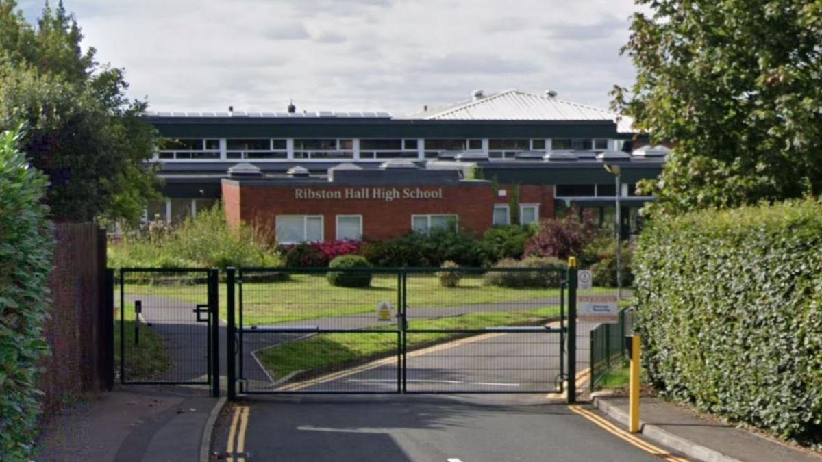A screenshot taken from Google street view showing the entrance to Ribston Hall High School. There is a green metal gate blocked the road entrance, and tall hedges on either side. The building itself is brown brick with a triangular white roof on the right.