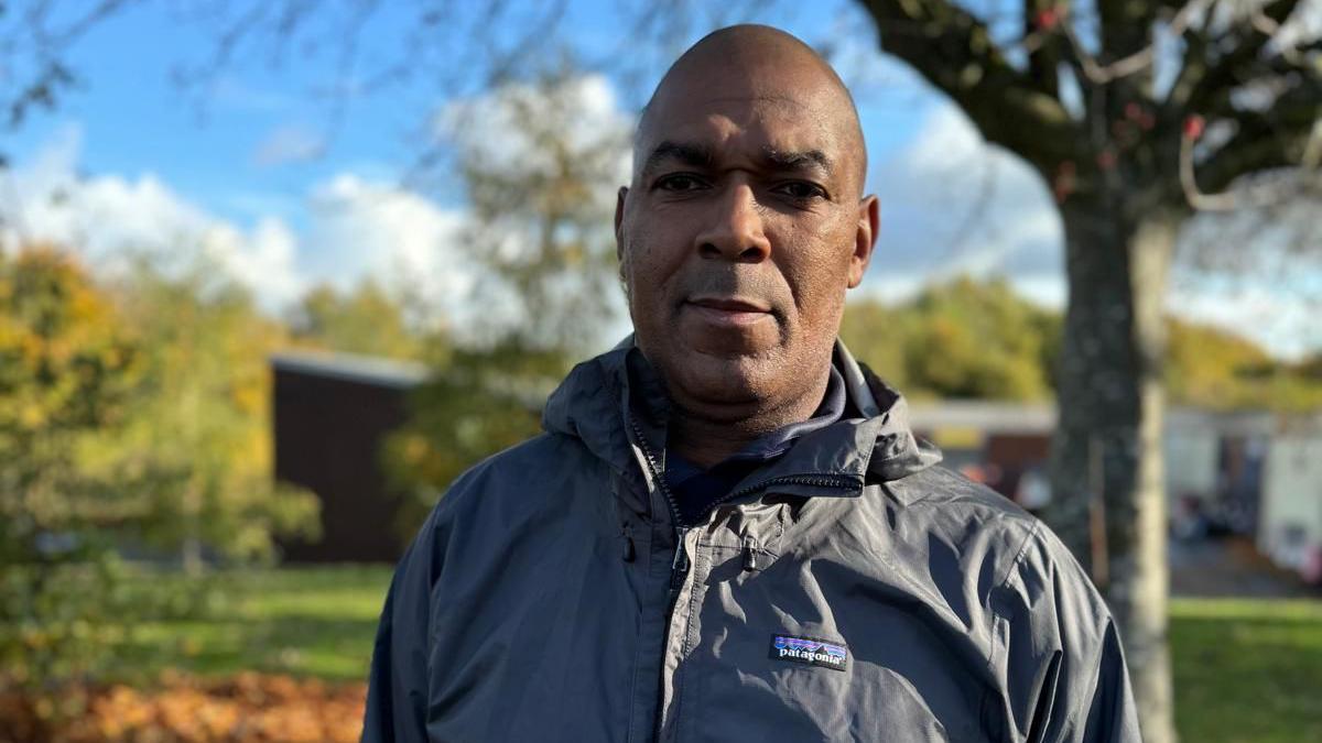 Rodney Tucker, wearing a grey Patagonia raincoat, looks into the camera with a serious expression against a backdrop of a grassy area and a tree