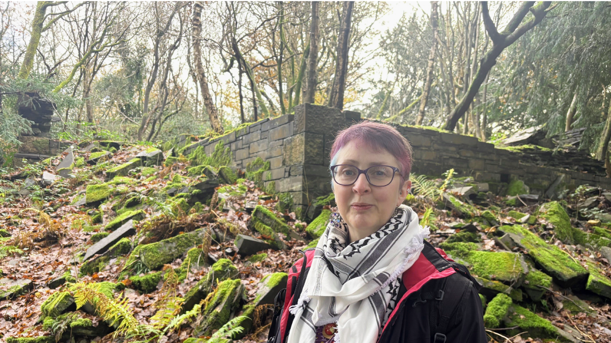 The Reverend Sara Roberts in a white and black scarf and black raincoat with pink trim stood in front of some moss covered gravestones that have fallen in every direction. She has pink, short hair with a blue streak on the left-hand side of her head and large purple-rimmed glasses 