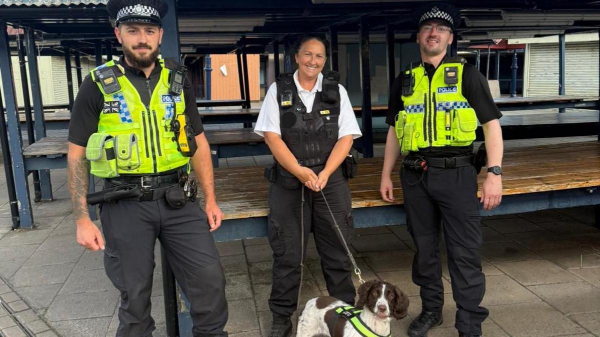 Two male police officers in black uniforms and yellow vests stand either side of a female officer holding a small dog on a leash in what appears to be an empty market area. She wears a black uniform over a white shirt. The dog is in front of her and is brown and white in colour with a yellow collar.