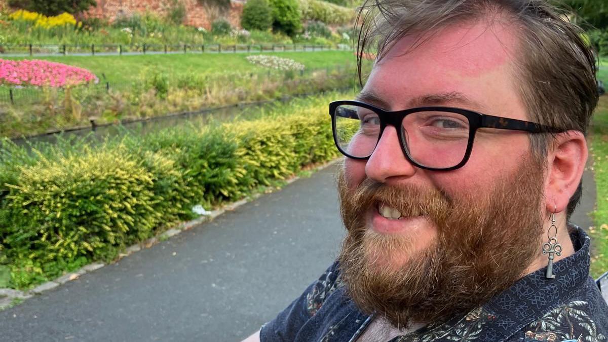 A man with a brown beard and glasses smiles at the camera while sitting in a park