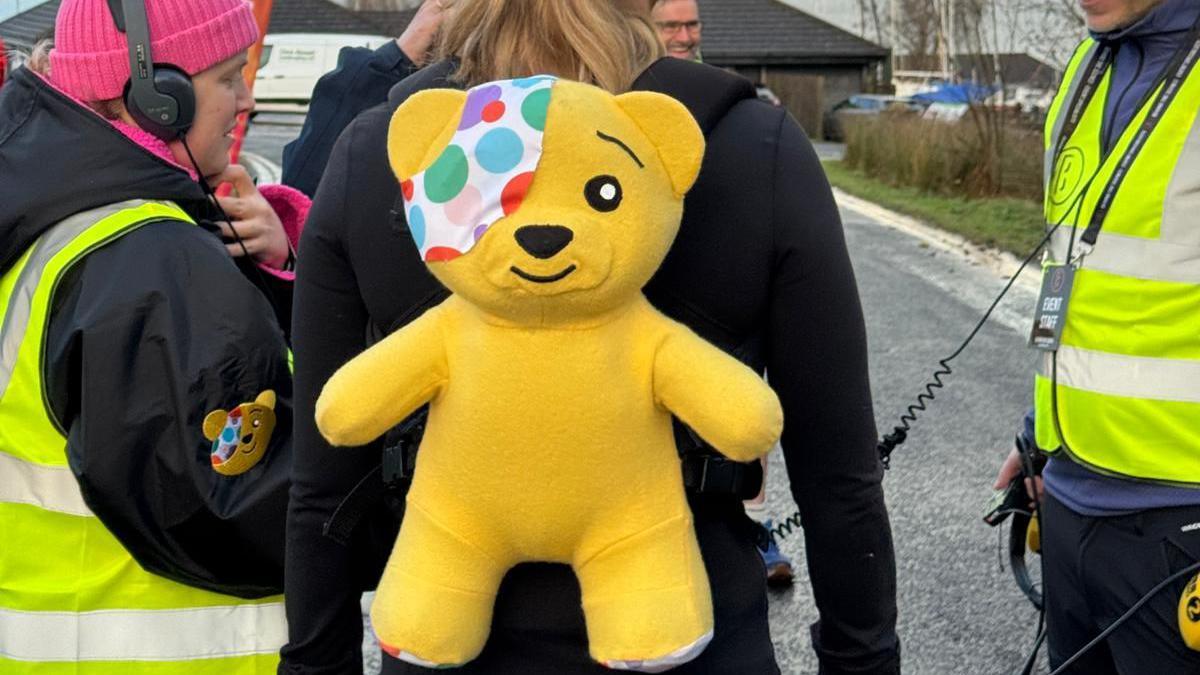 Sarah Cox wearing a black jumper stands facing away from the camera. She has a yellow Pudsey Bear soft toy strapped to her back. A woman wearing headphones stands to her left. 