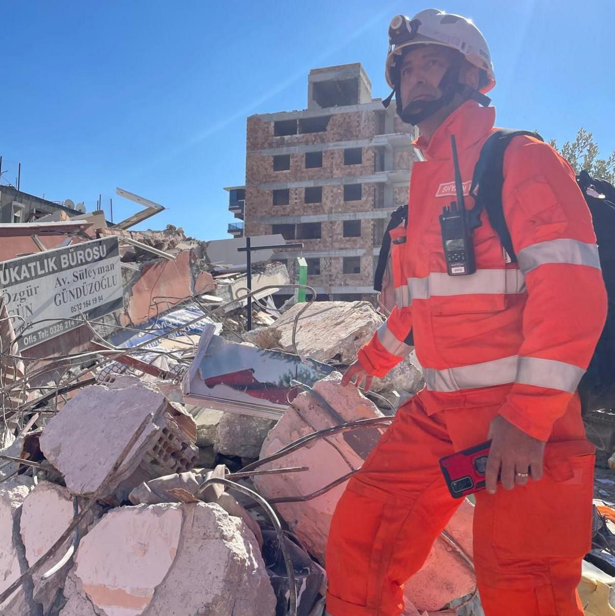 A man stands amongst a pile of rubble on a sunny day. The man is wearing a high-vis orange suit and a white helmet. A shell of a building with no glass in the windows can be seen in the background.