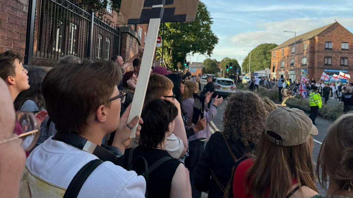 The two protests are taking place on opposite sides of the road, with police in hi-vis jackets in between. England flags are being held at the hotel demo and the counter-protesters have placards.