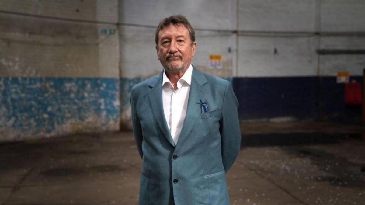 Peaky Blinders creator Steven Knight in a blue suit and a white shirt standing in a warehouse
