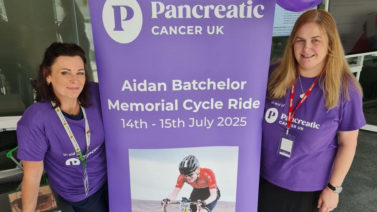 Two women dressed in purple T-shirts stand either side of a large purple banner with the words "Memorial Cycle Ride".