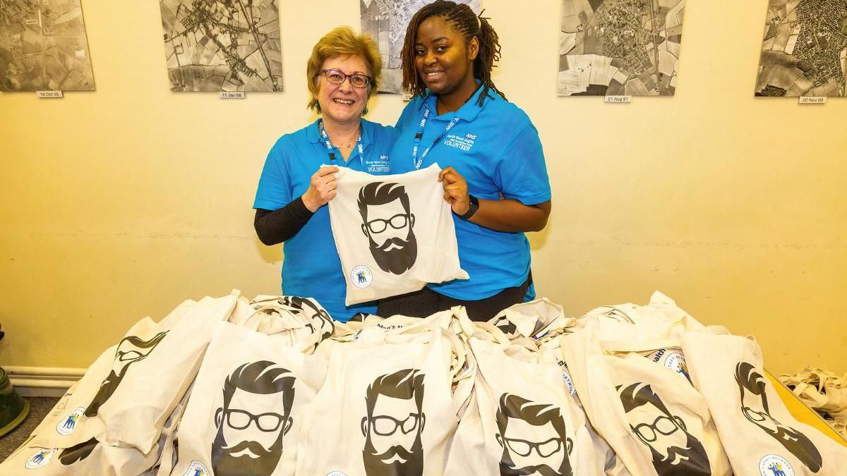 Two women holding a cloth bag with a male face sporting a beard and moustache. They are surrounded by bags with the same print on a table.