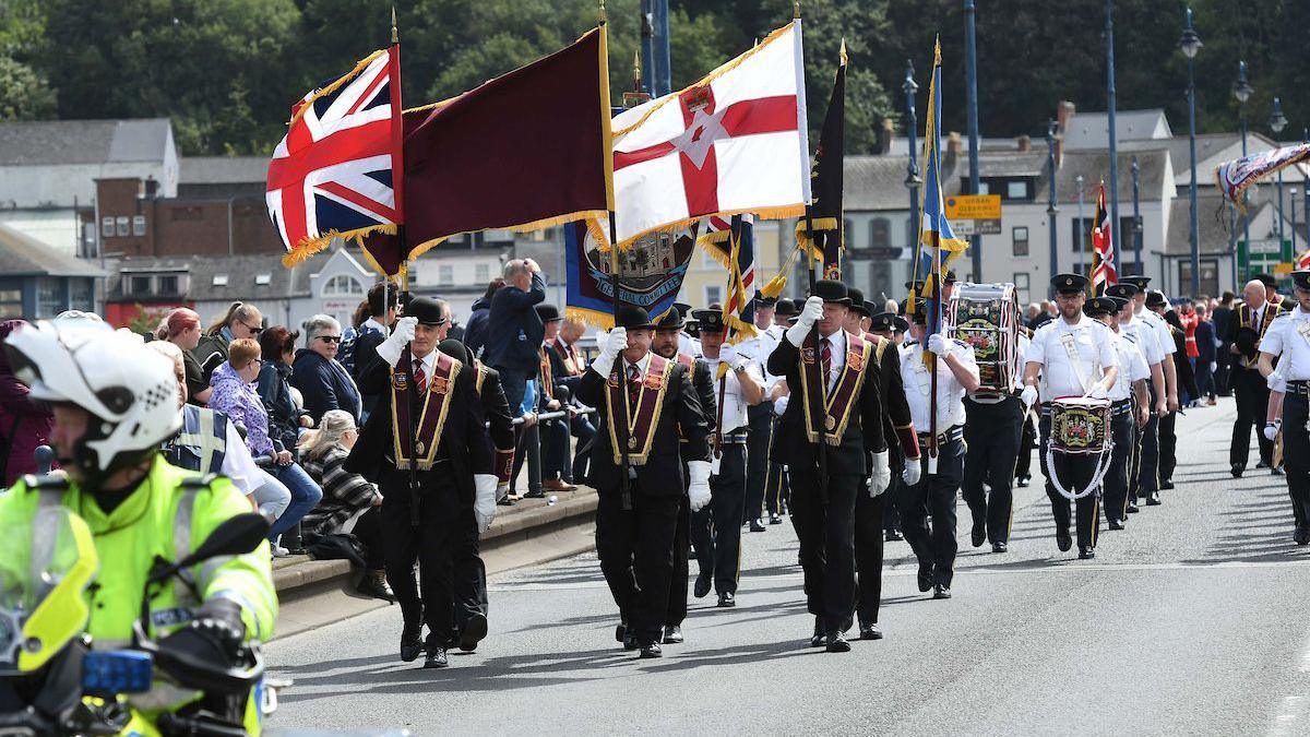 A number of men wearing suits with red ties, and ceremonial garments, marching in a parade. Some of them are holding flags. Behind them is a marching band. 